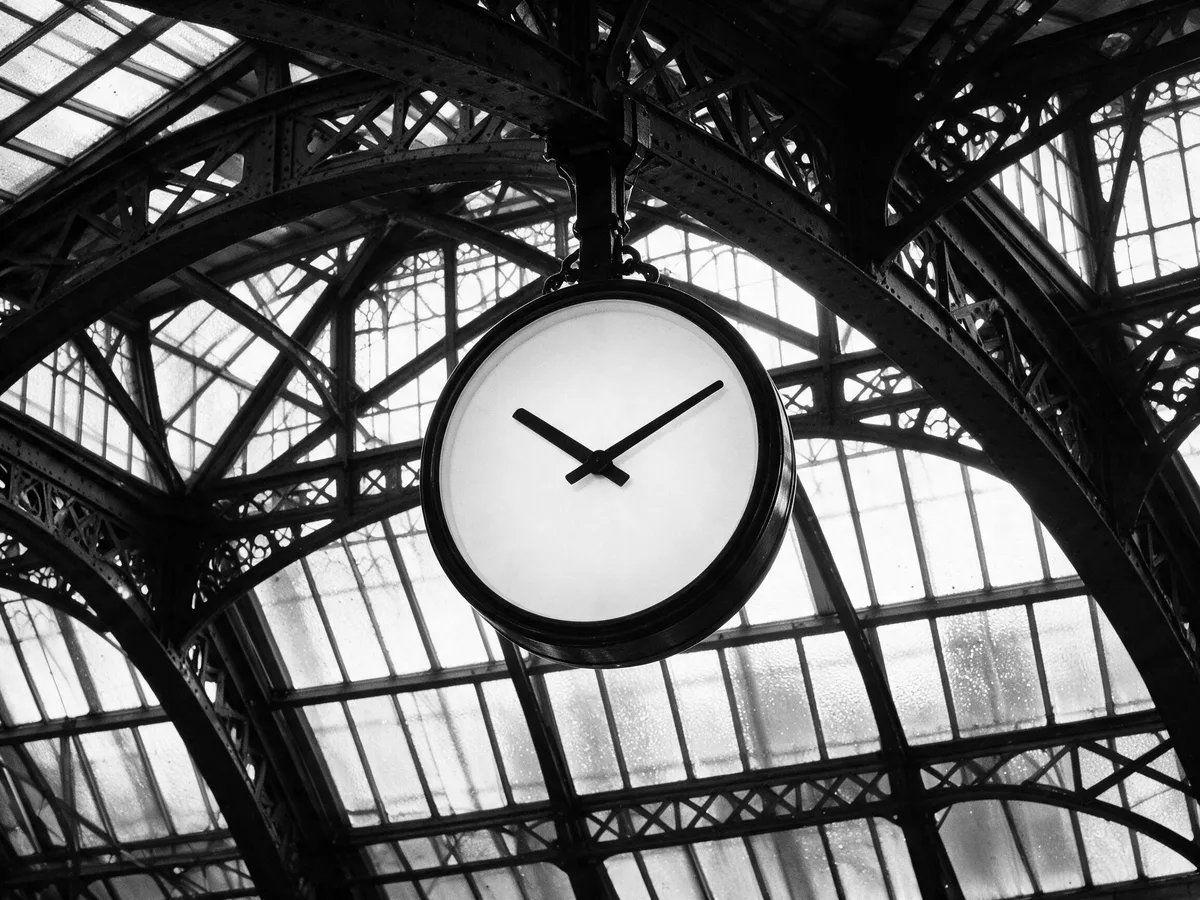A station clock hanging under the iron roof of a trainshed, in black and white.